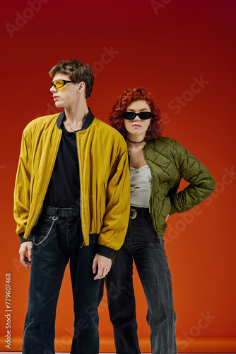 attractive stylish couple with cool sunglasses in cozy attires posing on red backdrop together