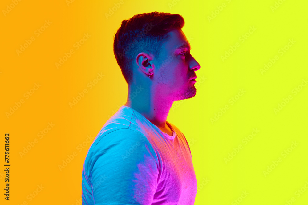Side view portrait of young handsome man posing in white T-shirt in neon light against vibrant yellow studio background. Concept of beauty, male health, anti-aging, spa procedures, cosmetic products.