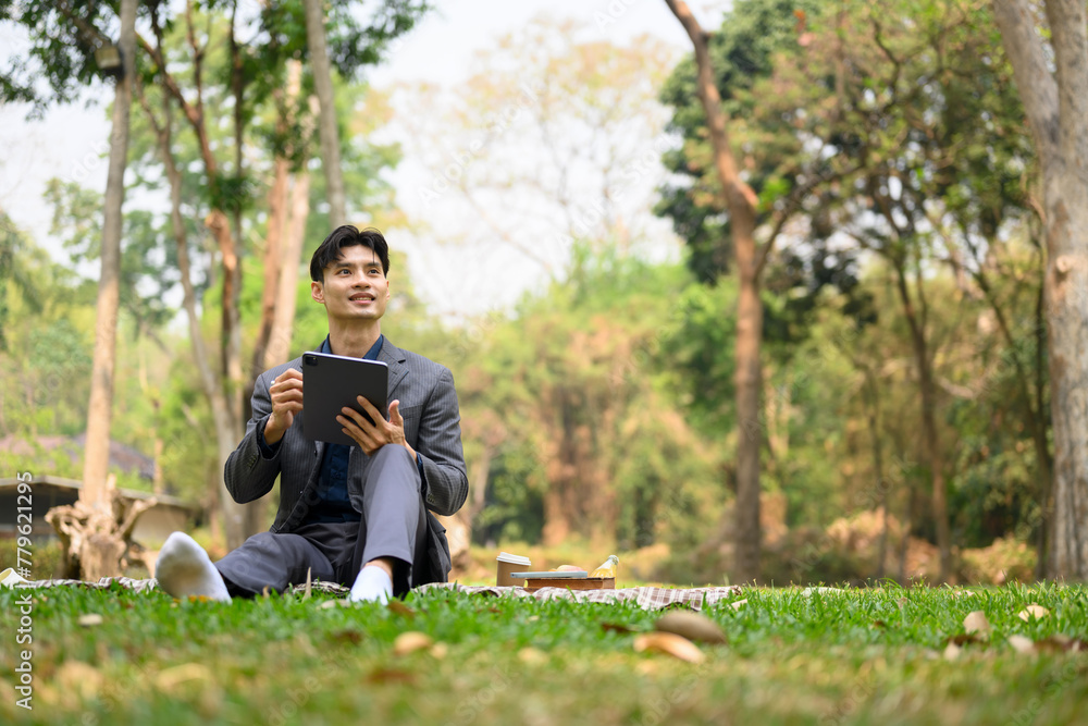 Carefree Asian businesswoman using digital tablet while sitting on grass in the summer park Carefree Asian businesswoman using digital tablet while sitting on grass in the summer park