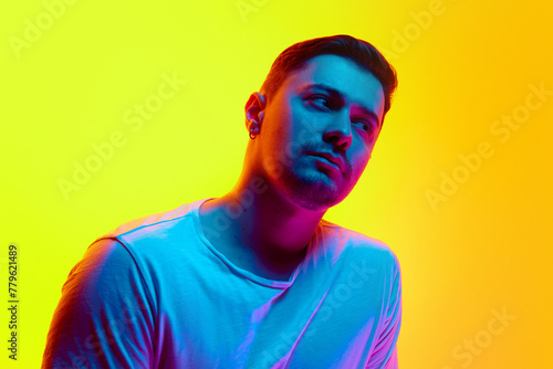Serious young athletic man posing in casual T-shirt in neon light against vibrant yellow studio background. Concept of natural beauty, male health, anti-aging, masculinity.