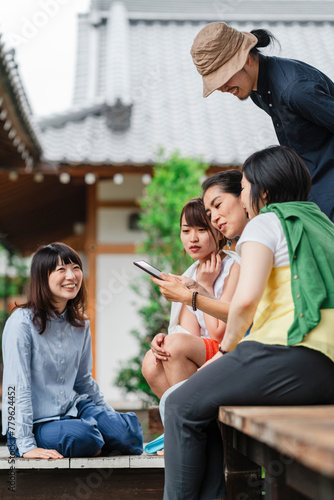 Five Japanese friends share a joyful moment looking at a tablet, laughter and smiles abound in an outdoor setting with traditional architecture in the background photo