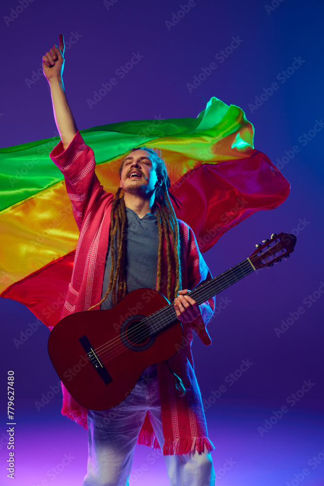 Fototapeta premium Energetic musician, man with striking dreadlocks playing guitar over red, yellow, and green flag against purple background in neon light. Concept of music, performance, festival, concert
