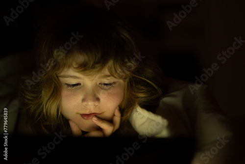 Cute little kid lying under blanket with tablet. Surprised kid in bed under blanket watching video on tablet in dark bedroom with glowing lights. Kid on bed at night watching movie.