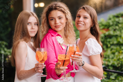 Three females hold alcoholic cocktails in hand and smile into camera. Hen tarty. Aperol sprits