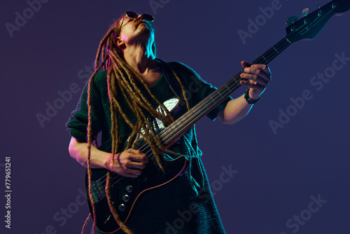 Charismatic guitarist with dreadlocks, musician emotionally playing electric guitar against dark purple background in neon light. Concept of music, performance, festival, concert, art