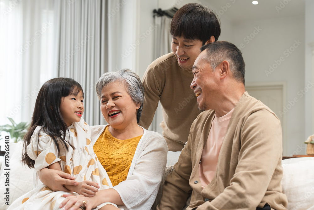Fototapeta premium Asian cute little girl sitting on sofa with grandparent and young father, talking, smiling together. Happy grandchild listen to grandfather fun story at home. Multi generation family relaxing together