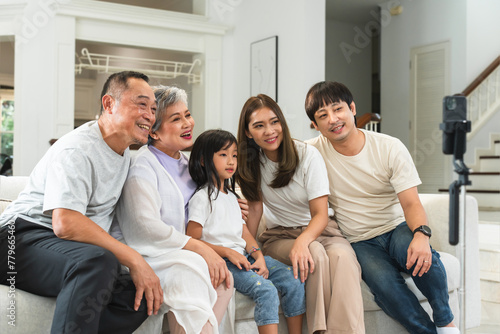 Happy Asian family, grandparent, mother, father and little cute daughter sitting on sofa together, taking photo with smartphone with happiness at home