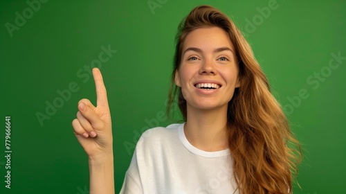 Portrait of beautiful happy American woman with long hair in white t-shirt pointing up and smiling at camera on green background, template for advertising or promotion, with copy space for text