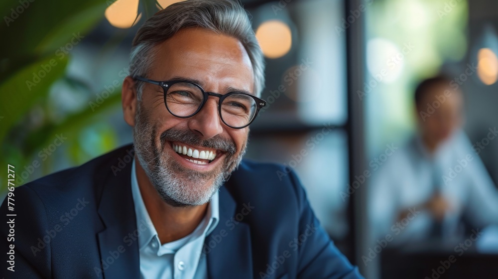 Happy bearded man smiling while sitting at a table in an office wearing glasses Happy bearded man smiling while sitting at a table in an office wearing glasses