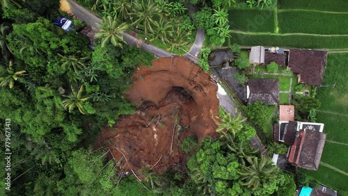 Top-down aerial shot of landslide aftermath, village road crossing destroyed, big mass of ground fall down. Unstable soil at steep slope of ravine was saturated by water from leaking subak channel