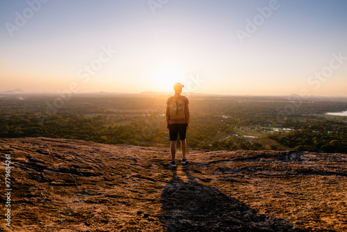 Rear view of man with backpack on rock overlooking sunset over tropical landscape. Travel in Sri Lanka..