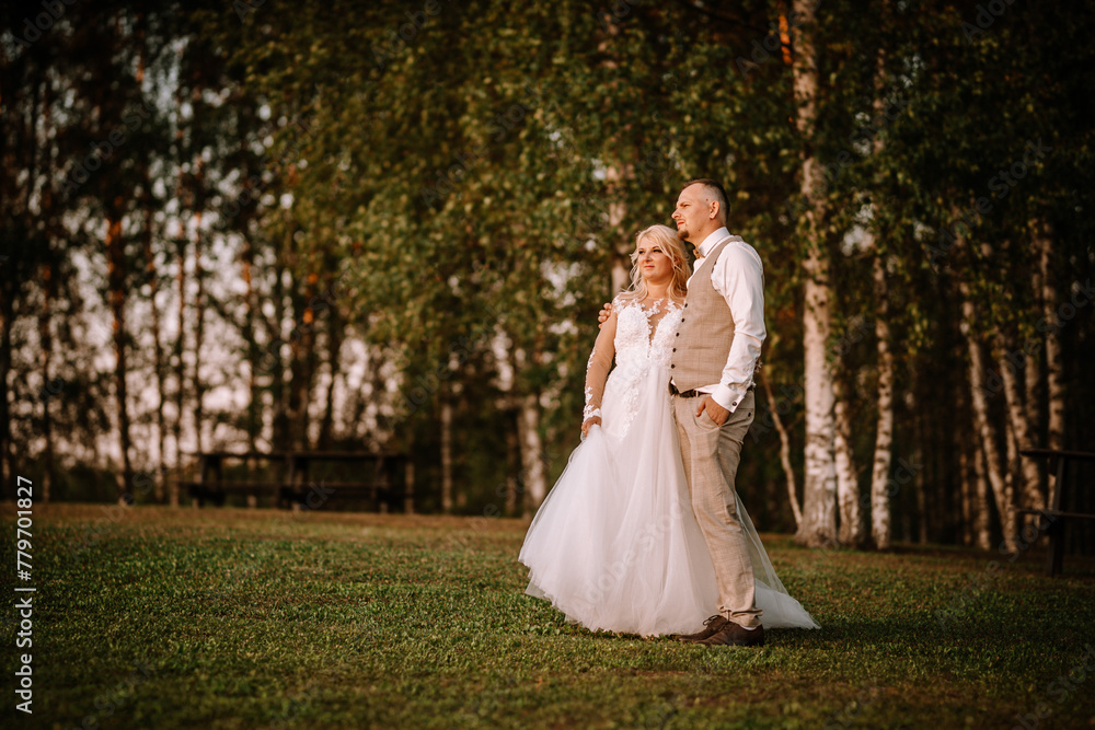Valmiera, Latvia - August 13, 2023 - A bride and groom stand side by side in a park, surrounded by trees, with the groom's hand around the bride.