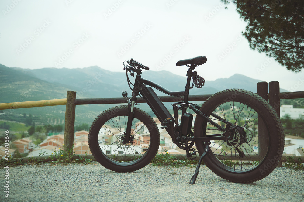 Fototapeta premium A full size shot of a modern black electric motor bike on the countryside road, parked by a wooden fence, against mountains background in the nature. Copy advertising space. Bike sharing city service.