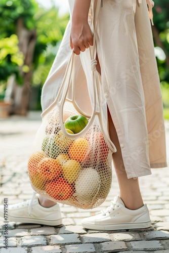 Fashionable woman holding an organic cotton net bag with fruit and vegetables, white sneakers, summer outfit, closeup on the shoes, blurred background of garden or terrace. Minimalistic concept.