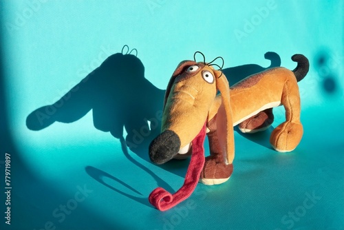 close-up of a looking into the lens with her mouth open and a very long tongue sticking out soft toy of a funny light brown dachshund on a blue background with a big shadow on the wall