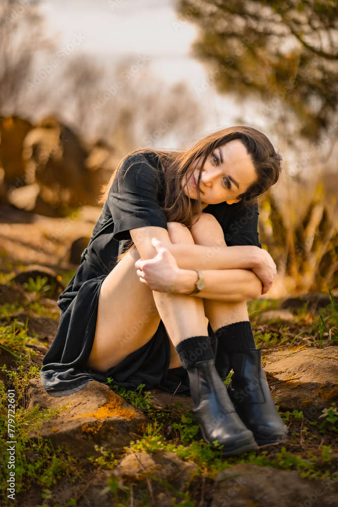 girl sits on rocks in a black dress, hugging her knees, against the backdrop of trees at sunset