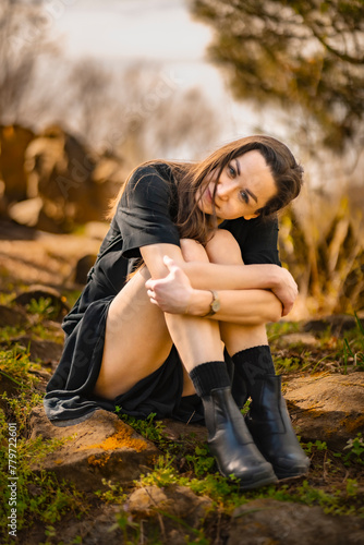 Obraz na płótnie girl sits on rocks in a black dress, hugging her knees, against the backdrop of trees at sunset w salonie