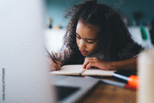 Serious African American curly haired female child studying and taking notes in notebook while sitting at wooden table with laptop against blurred background in living room photo