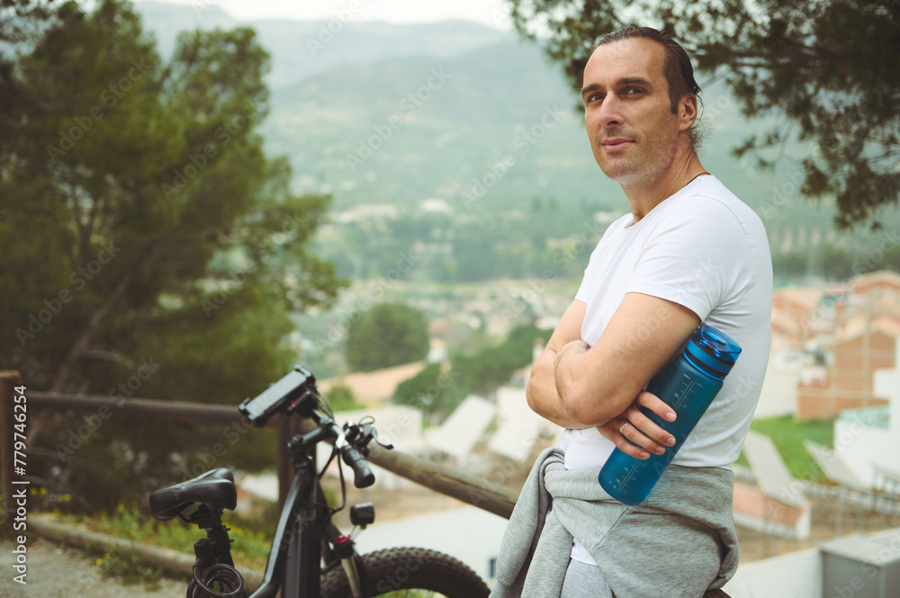 Handsome athletic man in white t-shirt, holding a bottle with water, smiling confidently looking at camera, relaxing after biking in mountains, standing by wooden fence near his electric bike