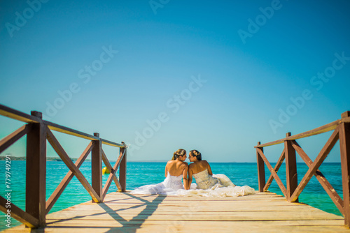 dos mujeres sentada mirandose frente a frente y usando vestidos de bodas en un muelle a la orilla de la playa 
