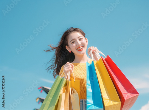 A beautiful young Asian woman is carrying colorful shopping bags and smiling happily, with a blue sky background behind her