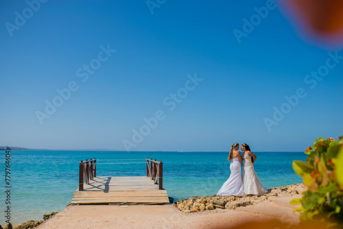 mujeres jovenes usando vestidos de boda y abrazadas mientras caminan junto a un muelle a la orilla de la playa 
