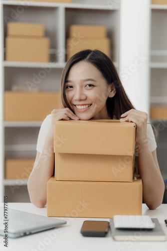 Freelance Asian woman wearing an apron uses laptop and boxes to take and check online orders to prepare packs to sell to customers. SME online business idea, vertical image