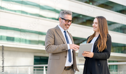 Experienced businessman with grey hair smiles as he looks at a tablet screen held by a young female professional, representing a merger of seasoned expertise and modern technology in corporate setting