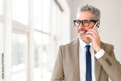 Cheerful senior 60s businessman talking on his smartphone, his pleasant demeanor reflecting a positive business discussion or negotiation, set in an airy, sunlit office.