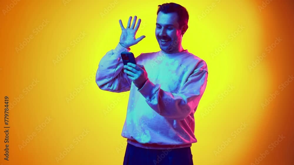 Young smiling man in sweatshirt and jeans having online video call via mobile phone on gradient yellow orange background in neon light. Concept of human emotions, casual fashion, lifestyle Young smiling man in sweatshirt and jeans having online video call via mobile phone on gradient yellow orange background in neon light. Concept of human emotions, casual fashion, lifestyle