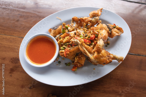 Thailand food - Fried Frog with Pepper and Garlic and fresh chilies on a white plate, served with Sriracha chili sauce in a small white bowl on a brown wooden table.