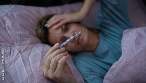 Photos Sick woman lying in bed and holding thermometer with high temperature closeup