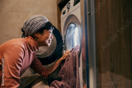 A happy japanese housewife is putting laundry into a washing machine in bathroom.