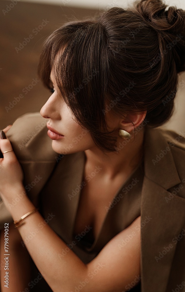 A young brunette woman with a sweet, kind face, dressed in a stylish brown suit with a jacket, trousers, and a vest, posing in a well-lit photo studio with a parquet floor.