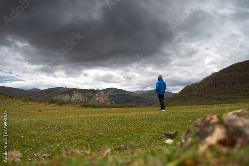 A man in a blue jacket stands in a field, surrounded by green grass and mountains with stormy sky