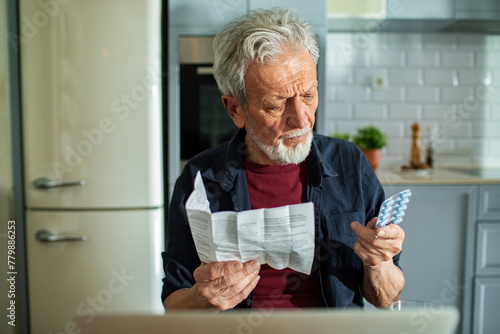 Senior man examining medication while holding documents at kitchen table with laptop and glass of water photo
