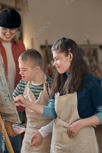 Vertical portrait of boy and girl with disability painting together on easel and smiling while enjoying art class