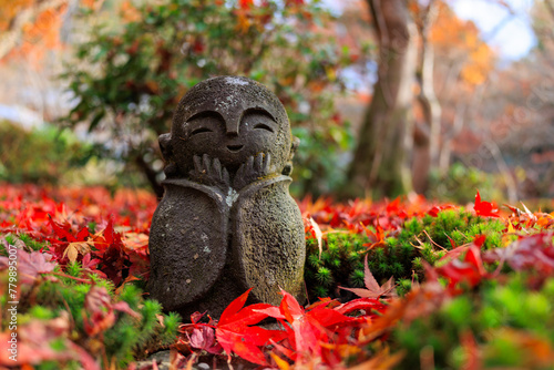 A Jizo statue with a smiling face is sitting on a pile of red leaves.