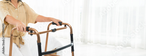 A woman in a tan shirt is using a walker. The walker is made of metal and has a black handle, panorama concept. © jittawit.21