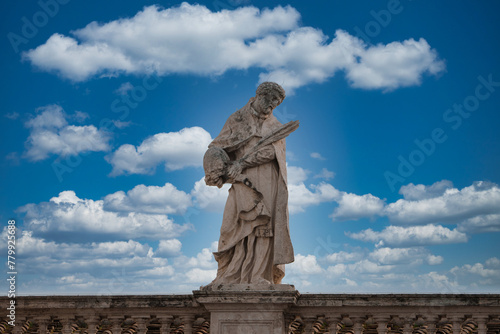 Stone statue of religious figure holding book and key against blue sky in Vatican. Weathered, with decorative balustrade. Rich collection of Vatican art.