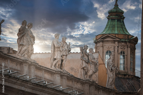 Classical marble statues under dramatic sky in Vatican City. Intricate carvings with religious or historical motifs. Dome topped structure nearby. Ethereal atmosphere with serene lighting.