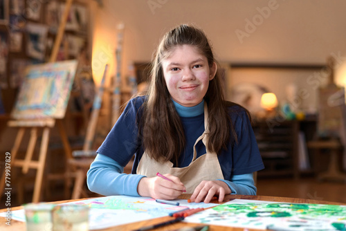 Front view portrait of young girl with Williams syndrome smiling at camera sitting at table in art studio