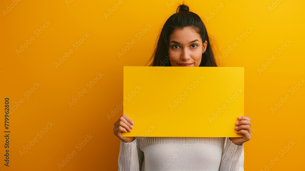 A woman is holding a yellow sign in front of a yellow wall. Concept of playfulness and creativity, as the woman is holding up a blank sign. Young woman hiding face with blank empty banner, mock up A woman is holding a yellow sign in front of a yellow wall. Concept of playfulness and creativity, as the woman is holding up a blank sign. Young woman hiding face with blank empty banner, mock up