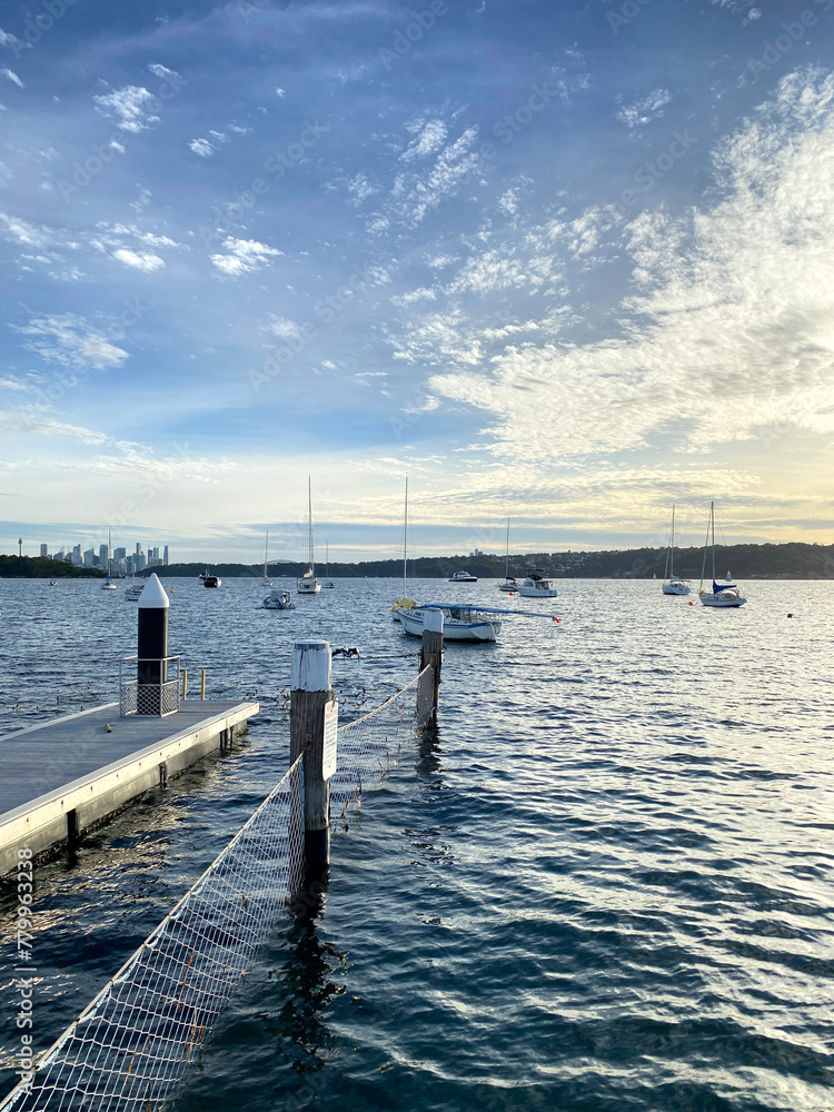 Pier in the sea. Lake with boats sailing. Wooden dock on the shore. Bollard for mooring boats. City of Sydney at the distance.