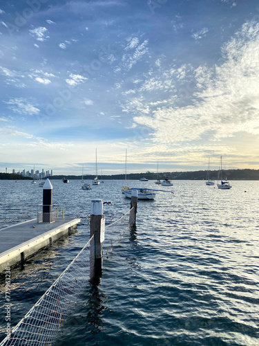 Pier in the sea. Lake with boats sailing. Wooden dock on the shore. Bollard for mooring boats. City of Sydney at the distance.