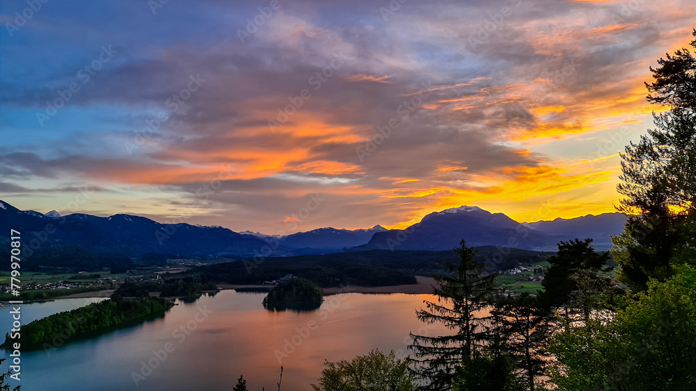 Naklejka premium Panoramic sunset view on Lake Faak from Taborhoehe in Carinthia, Austria, Europe. Surrounded by high Austrian Alps mountains. Water surface reflecting soft sunlight. Remote alpine landscape in summer