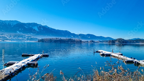 Snow covered wooden pier on lake Faak in Carinthia, Austria, Europe. Surrounded by high snow capped Austrian Alps mountains. Calm water surface with reflections of landscape. Looking at Dobratsch peak photo