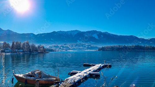 Snow covered wooden pier on lake Faak in Carinthia, Austria, Europe. Surrounded by high snow capped Austrian Alps mountains. Calm water surface with reflections of landscape. Looking at Dobratsch peak
