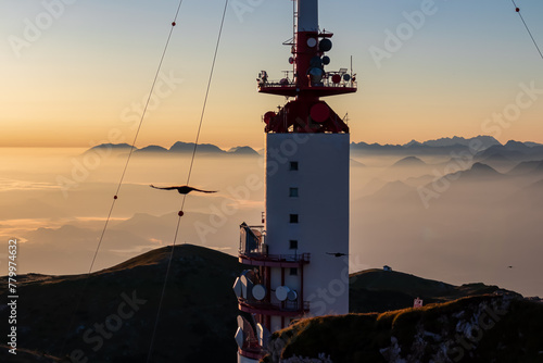 Birds flying around the radio tower with panoramic sunrise view from Dobratsch on Julian Alps and Karawanks in Austria, Europe. Silhouette of endless mountain ranges covered by mystical fog in valley
