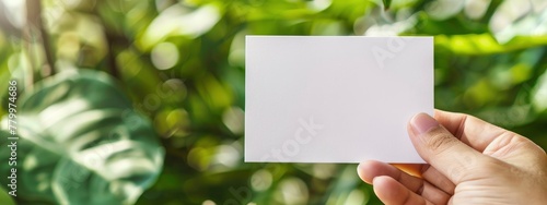 Closeup hand holding blank white card mockup on blurred green plant background, interior design with copy space area for text